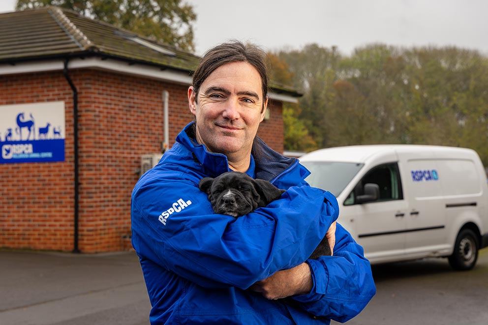 RSPCA worker in blue coat holds small black puppy in front of a white RSPCA van