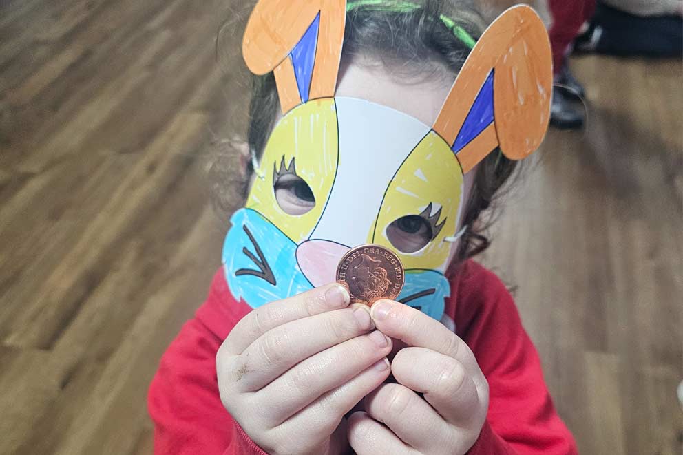 A child wearing a handmade paper bunny mask holding up a shiny 2p coin.